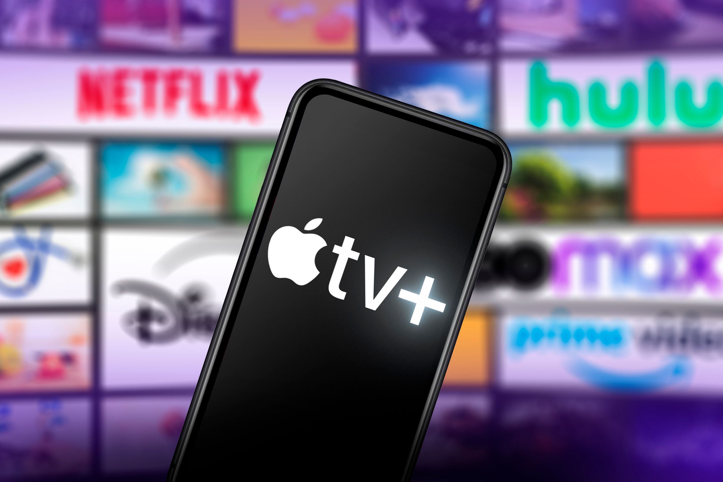 Một chiếc điện thoại có logo Apple TV+ trên màn hình và ở phía sau là một số màn hình có logo của các dịch vụ phát trực tuyến khác.